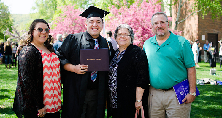 WSU student in graduation cap and gown stands with family while holding diploma outside on campus