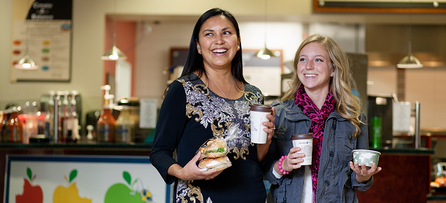 Two female students holding sandwhiches, coffee and soup in dining area